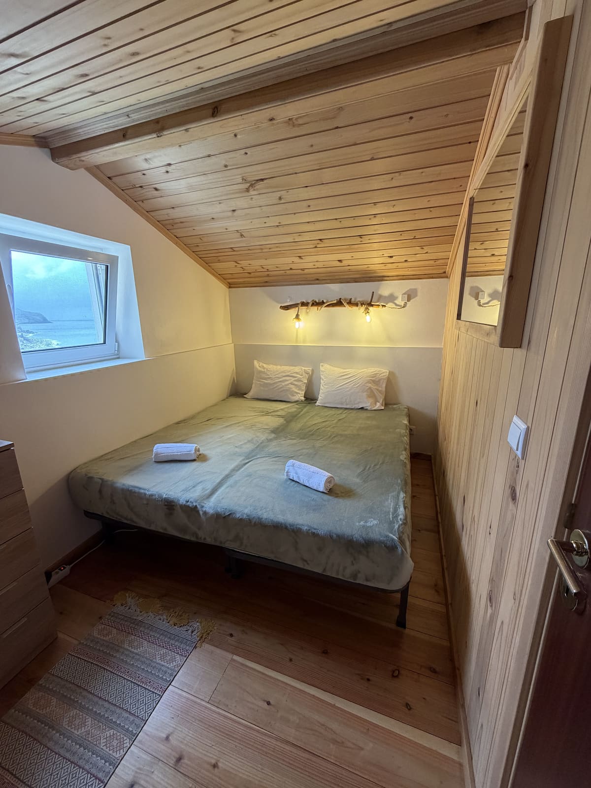 Loft bedroom with a double bed under a sloped wood ceiling, ocean visible through the window