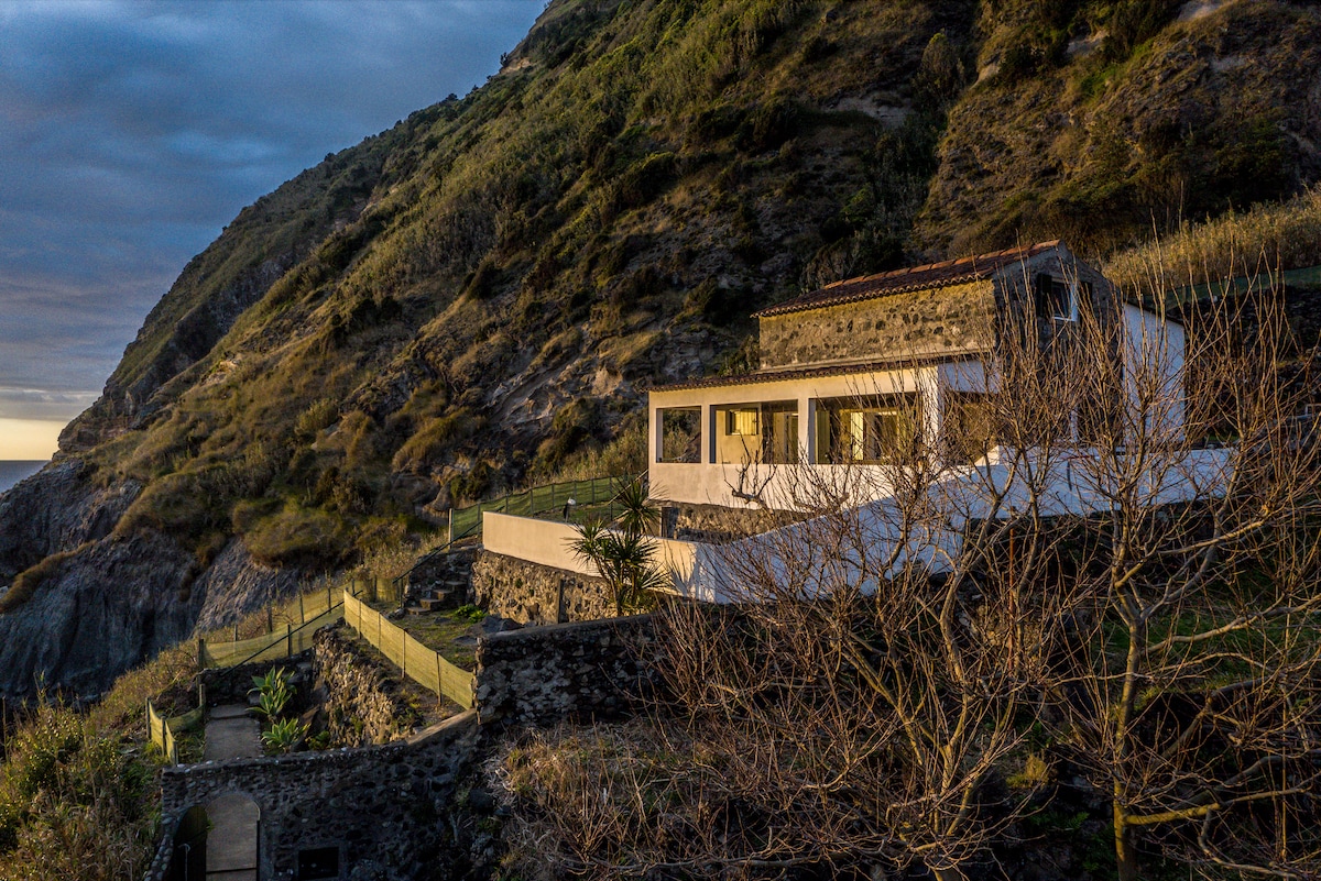 The house at golden hour, built into the cliffside above the Atlantic, terracotta roof and white walls against the mountain