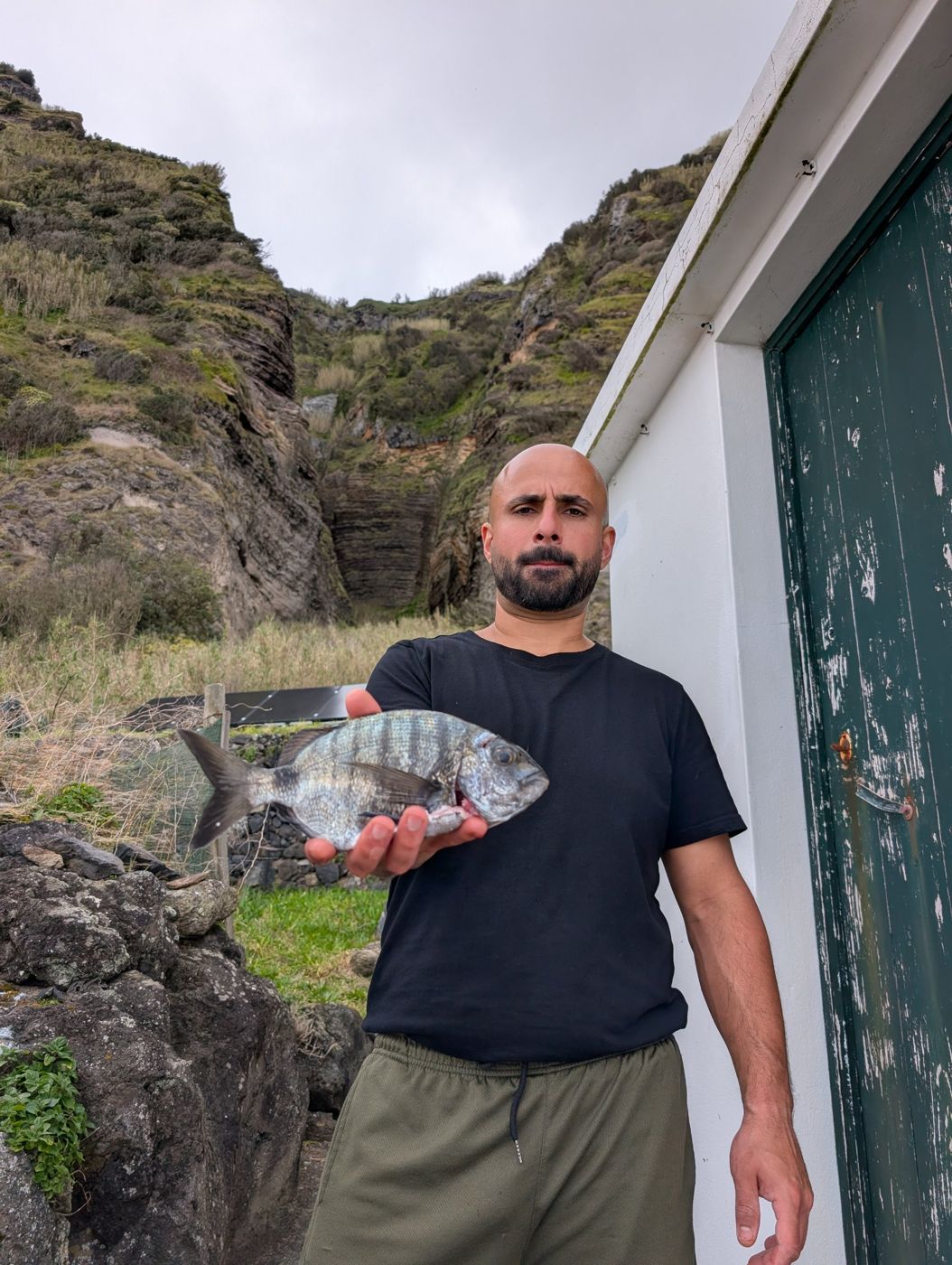 Ramsen holding a fresh sea bream by the green door, cliffs behind