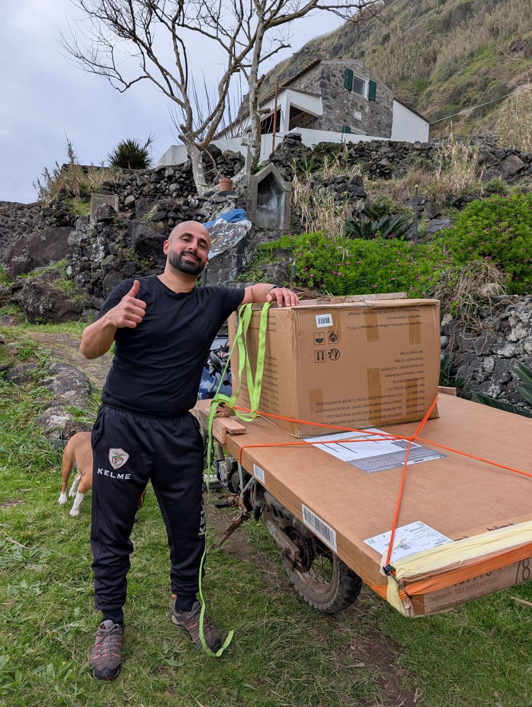 Ramsen giving a thumbs up beside furniture boxes strapped to a motorbike, the house behind
