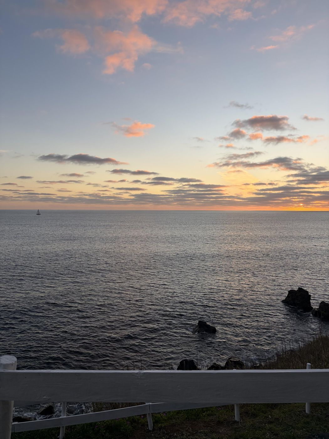 Sunset over the Atlantic with a single sailboat on the horizon, white fence in front